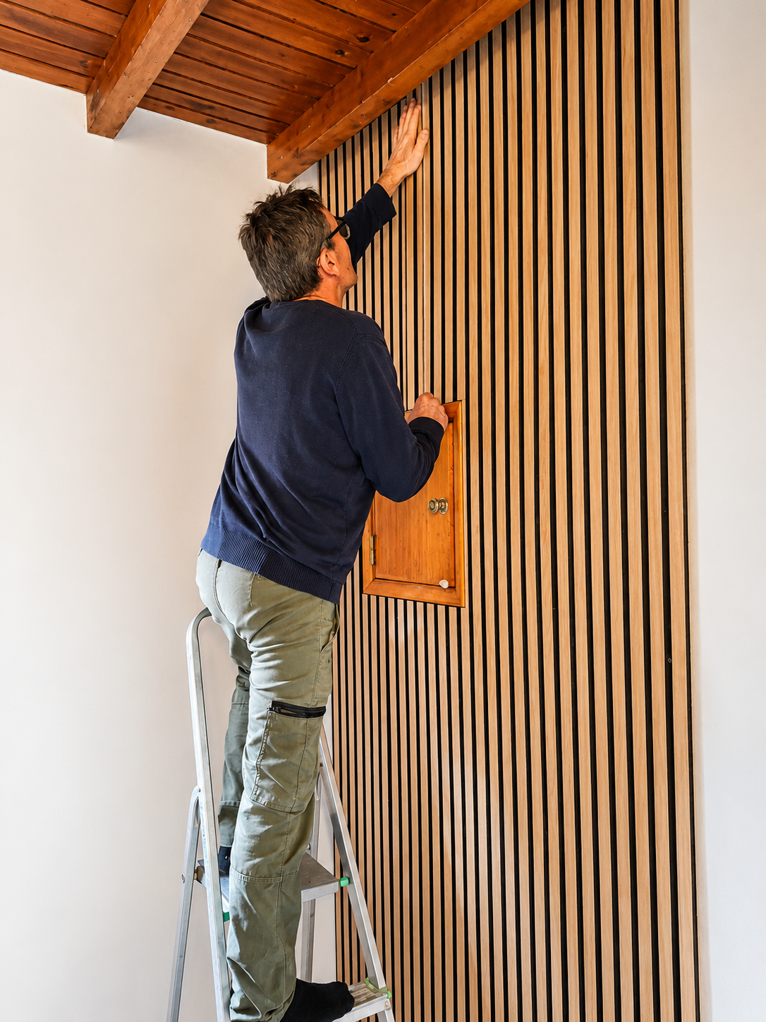 Handyman on a ladder finishing a wood slat wall with matching cabinetry